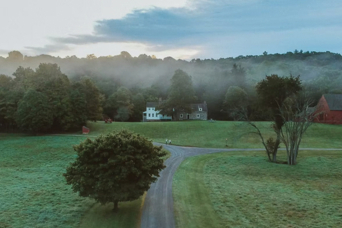 An aerial view of a sprawling historic farm estate nestled among rolling, verdant hills and mature trees under a clear sky.