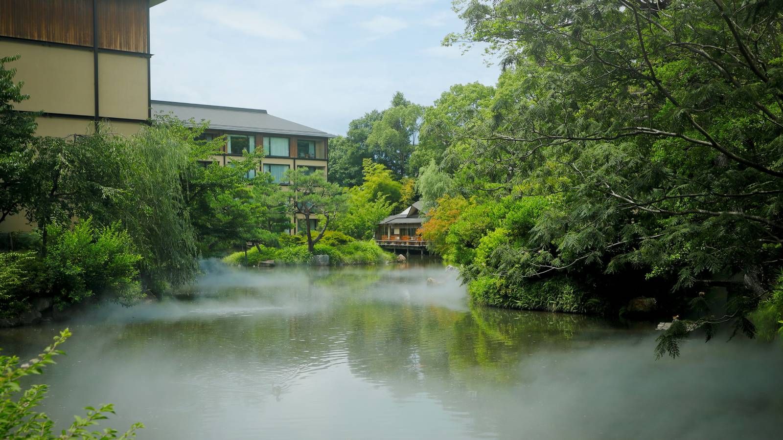 Vue d'un jardin-étang japonais ancien avec des arbres méticuleusement taillés et un pavillon traditionnel, encadré par des éléments architecturaux modernes du Four Seasons Kyoto.