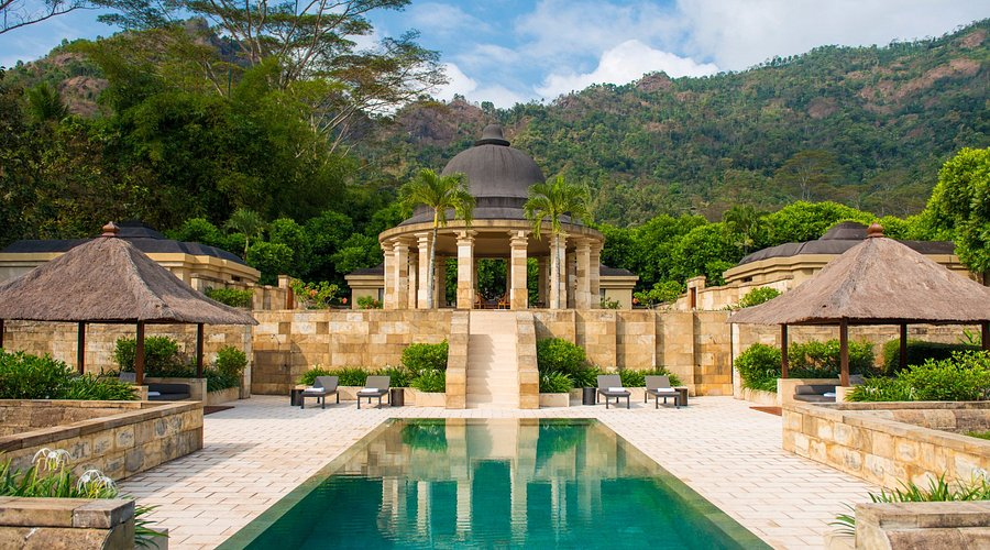 A woman practices yoga on a mat overlooking a serene outdoor pool and a lush landscape, suggesting a luxury resort setting.