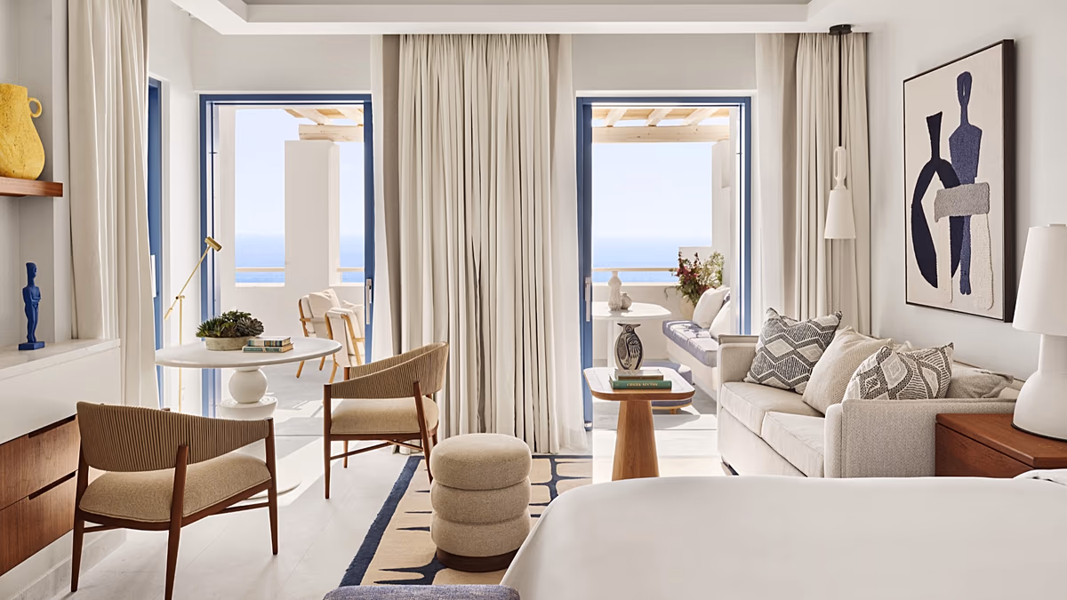 A luxurious resort with multiple pools and white buildings cascading down a cliffside to a private beach overlooking the Aegean Sea.