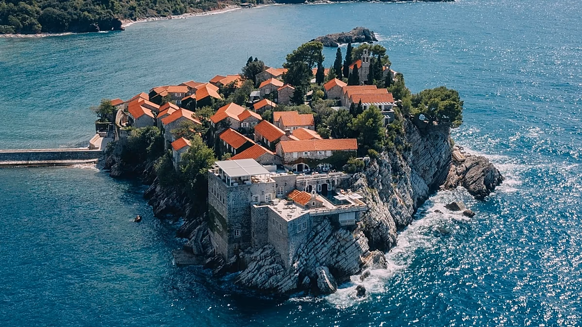 Ariel view of Aman Sveti Stefan, a fortified island resort connected to the mainland by a narrow causeway, with clear blue waters surrounding it.