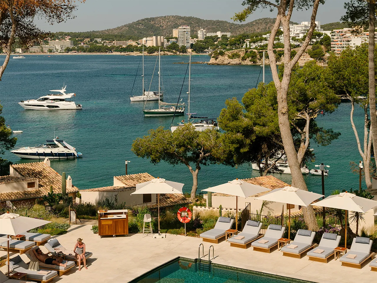 Aerial view of a luxury hotel complex nestled on a rocky coastline with pine trees, overlooking the Mediterranean Sea at sunrise.