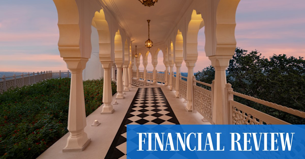 Exterior view of The Oberoi Rajgarh Palace, a grand edifice nestled amidst lush green hills and trees under a clear sky.