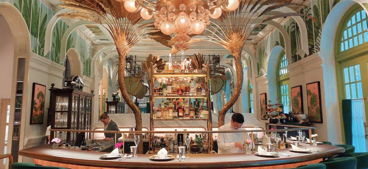 A grand bar area with metallic sculptures resembling palm trees, under a hand-painted azure ceiling, with a dramatic chandelier.