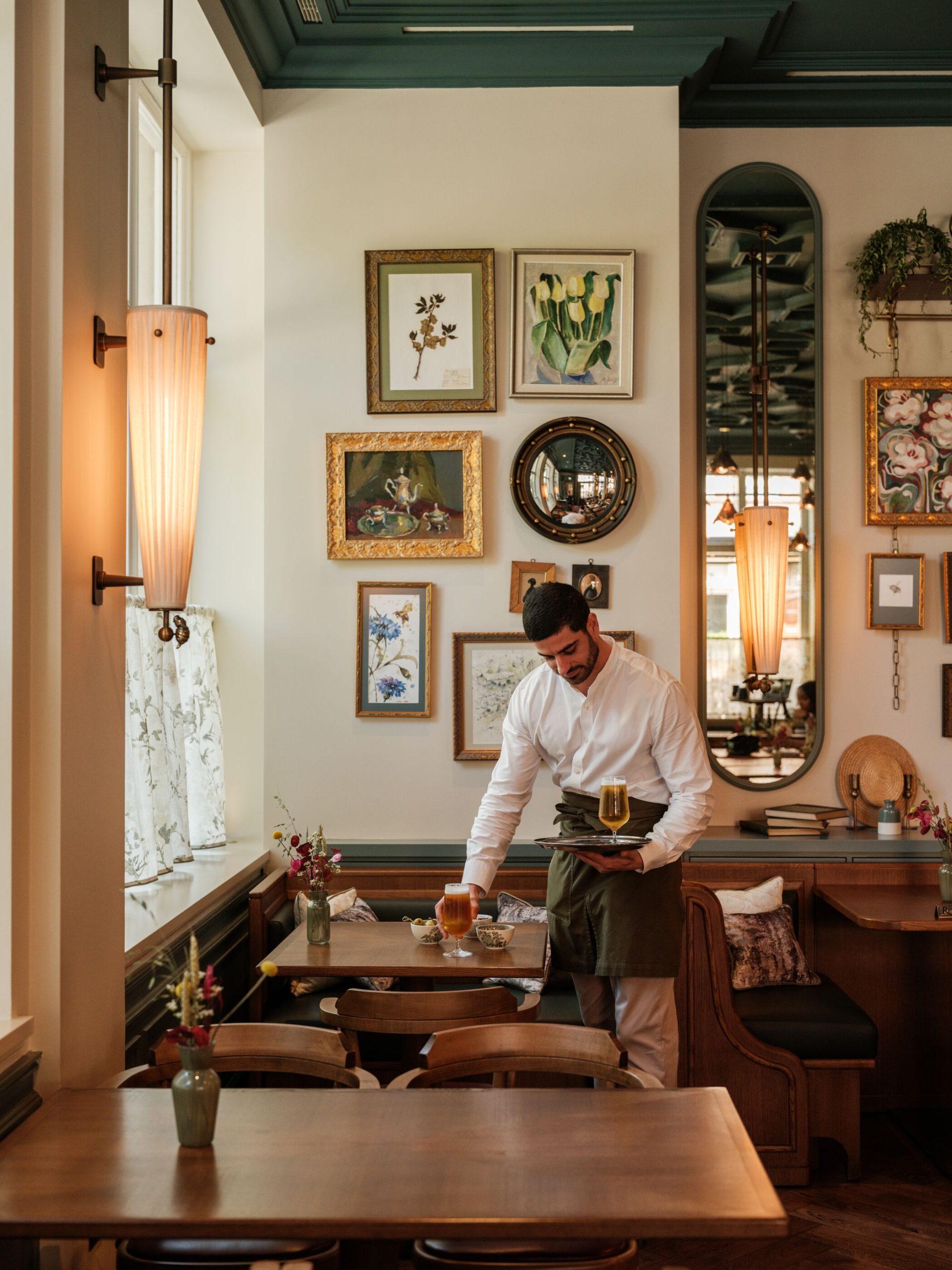 A stylishly lit bar area at Willett's at Belmond Cadogan with a person seated facing the camera, tables and stools, and bottles on shelves behind the bar.