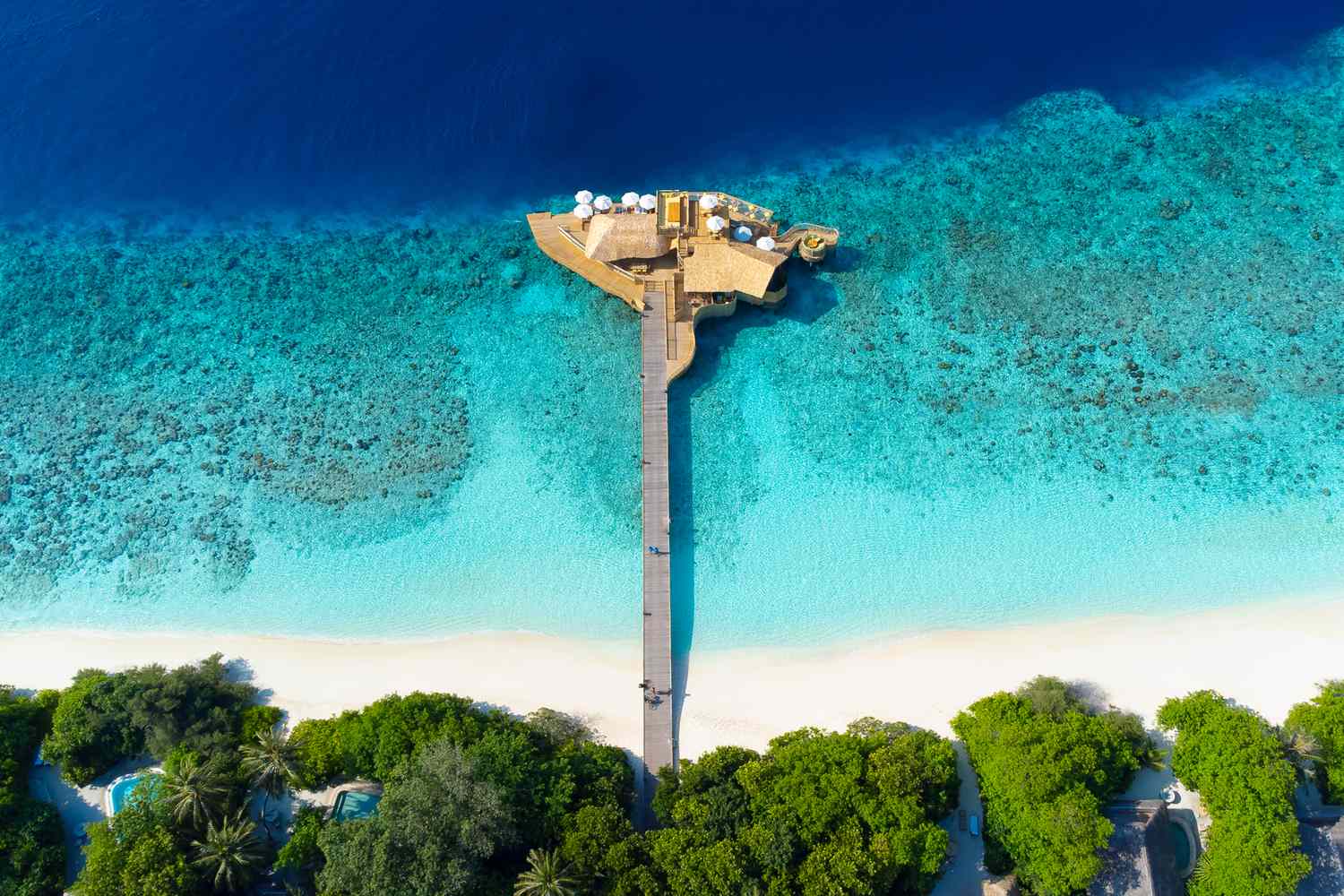Aerial view of a wooden pier extending over turquoise waters, connecting to a lush green beach